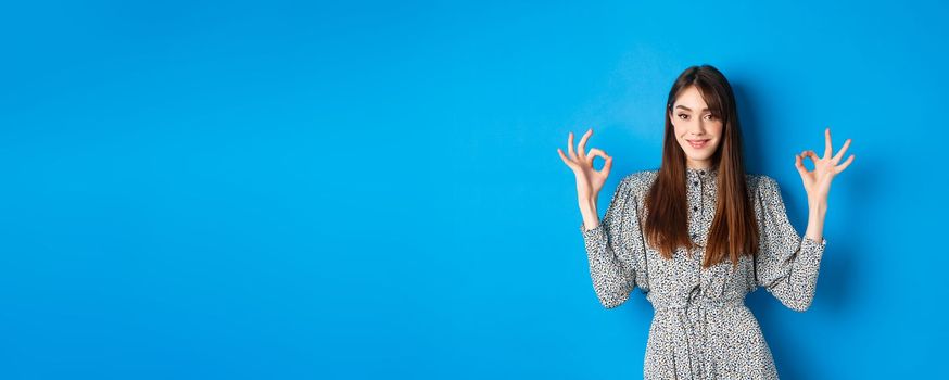 Confident adult woman in dress with natural hair color, showing okay signs and smiling, like and approve something good, standing on blue background.