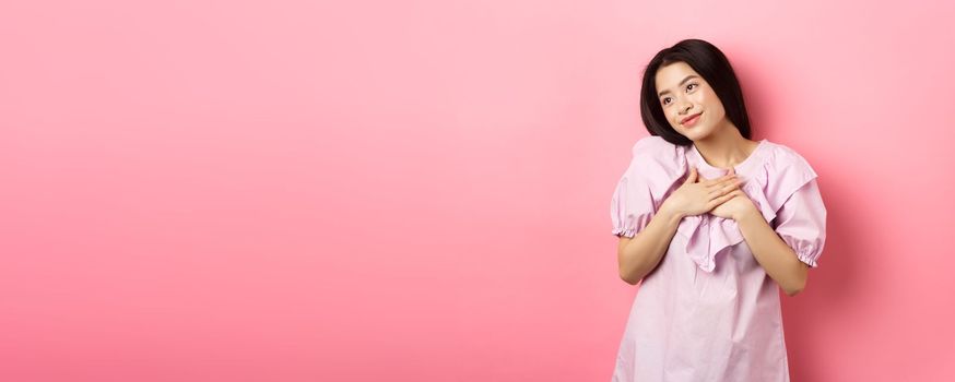 Dreamy asian girl smiling and holding hands on chest, looking left at something romantic and cute, standing on pink background.