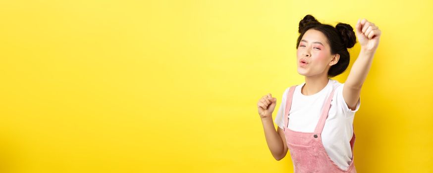 Excited asian girl looking motivated, raising hand up and chanting, cheering with joy, standing happy on yellow background.