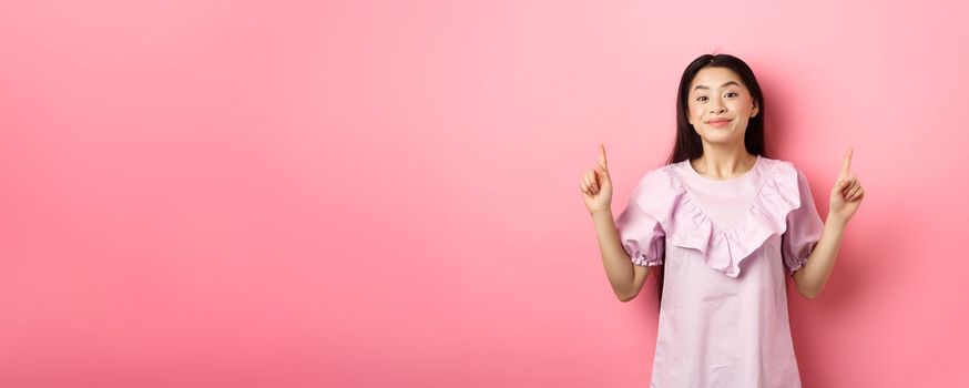 Beautiful young asian woman in romantic dress pointing fingers up, smiling and gladly showing advertisement, standing on pink background.