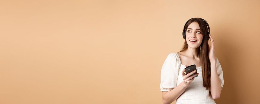 Attractive girl listening music in wireless headphones, holding mobile phone and looking aside with dreamy smile, beige background.