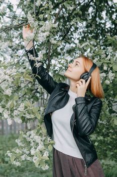 A red-haired teenage girl listens to music in headphones, configured via the phone. The time of spring flowering of cherry orchards.