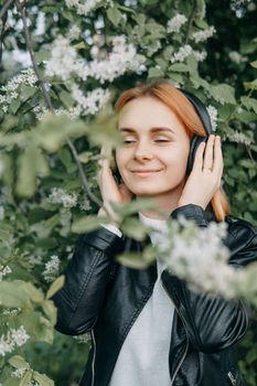 A red-haired teenage girl listens to music in headphones, configured via the phone. The time of spring flowering of cherry orchards.