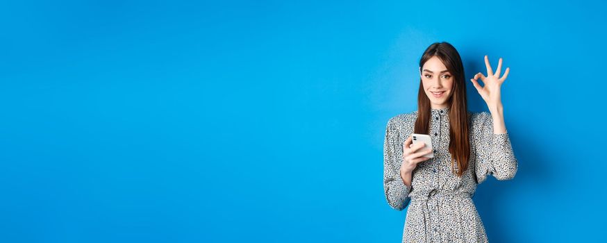 Beautiful young woman in dress showing okay sign and using smartphone, smiling at camera, standing on blue background.