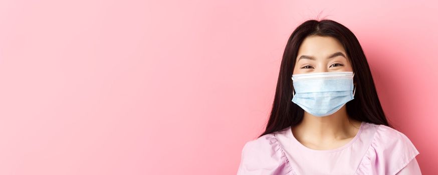 Covid-19 and healthy people concept. Close-up of cheerful asian teen girl wearing medical mask and smiling with eyes, standing against pink background.