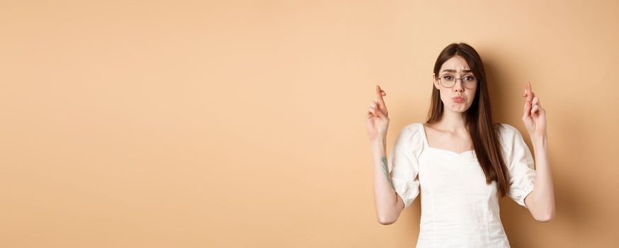 Cute hopeful girl making wish, pucker lips and begging, say please, standing on beige background.