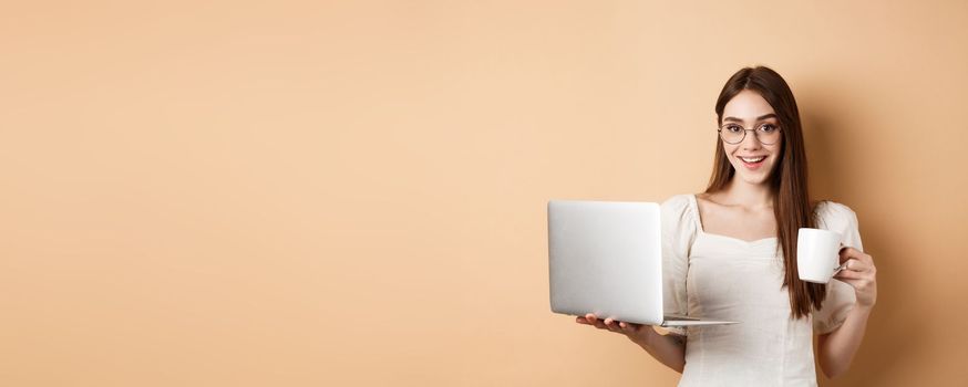 Cheerful woman in glasses drinking coffee and working on laptop, smiling at camera, standing on beige background.