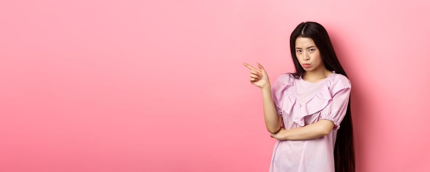 Sad and grumpy asian girl sulking, pointing left and look at camera jealous, want something, complaining and showing desired thing, standing on pink background.