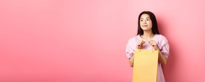 Beautiful asian girl dreaming and holding shopping bag, standing on pink background.