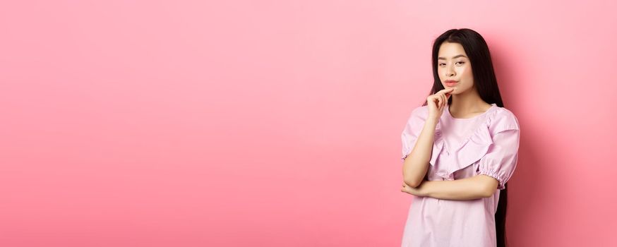 Hmm, interesting. Pensive and cunning asian girl having idea, plan to do something, looking at camera and touching chin with thoughtful face, standing on pink background.