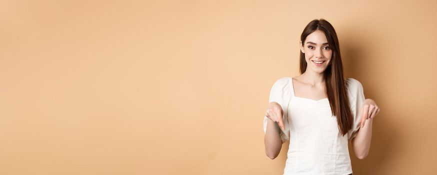 Cheerful young girl with lovely smile, pointing fingers down, making announcement or inviting click link, showing logo, beige background.