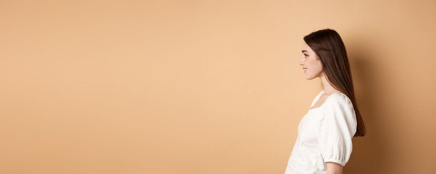 Profile of beautiful caucasian woman with long natural hair, looking left at empty space and smiling romantic, standing on beige background.