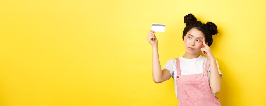 Shopping. Thoughtful trendy girl with summer makeup, looking pensive at plastic credit card, standing on yellow background.