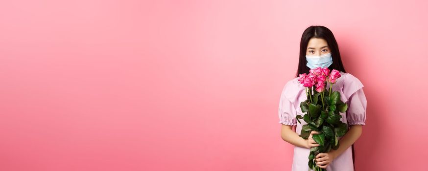 Young asian girl in medical mask holding flowers on Valentines day, receive bouquet of roses from lover, standing on pink background. Social distancing and covid-19 concept.