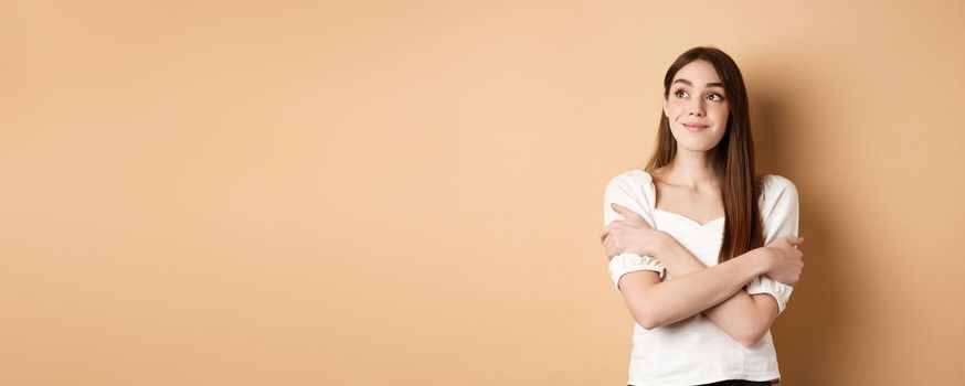 Romantic young girl dreaming, hugging herself and smiling at upper left corner, imaging something lovely, standing on beige background.