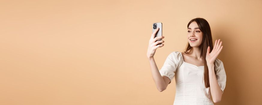 Girl video chat on phone, waiving hand at mobile camera and saying hello, talking to friend, smiling and standing on beige background.