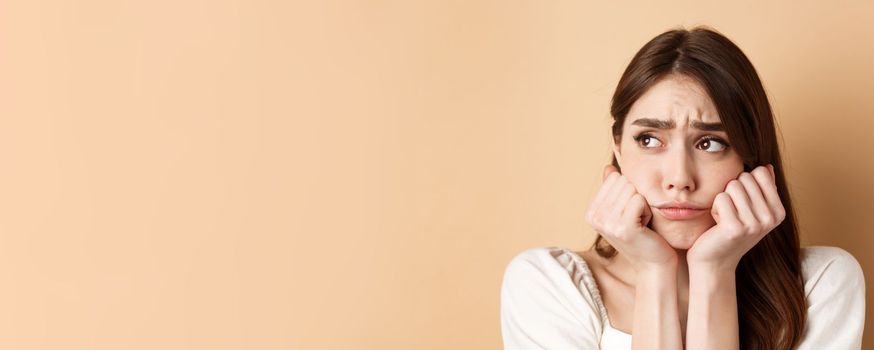 Close up of sad and gloomy girl looking aside, sulking and frowning upset, standing miserable on beige background.