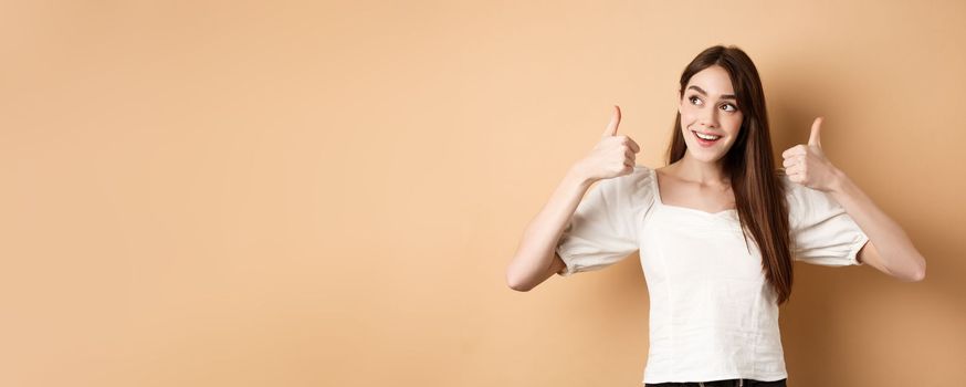 Smiling dreamy girl looking at upper left corner and showing thumbs up at promo, recommending product, praising banner, standing on beige background.