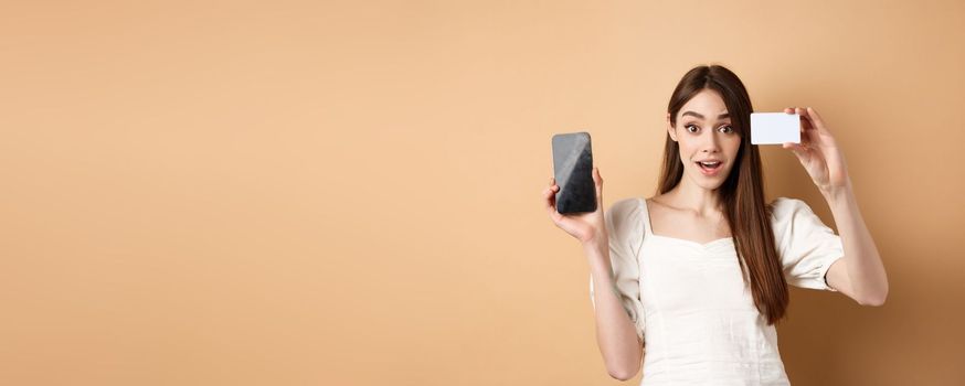 Excited girl showing plastic credit card of bank and empty mobile phone screen, demonstrate shopping app, standing on beige background.