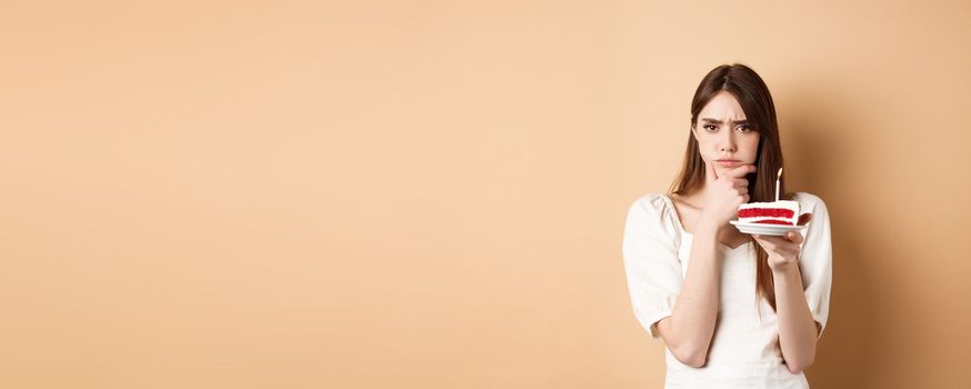 Thoughtful birthday girl frowning and thinking of wish, holding cake with candle, standing on beige background.