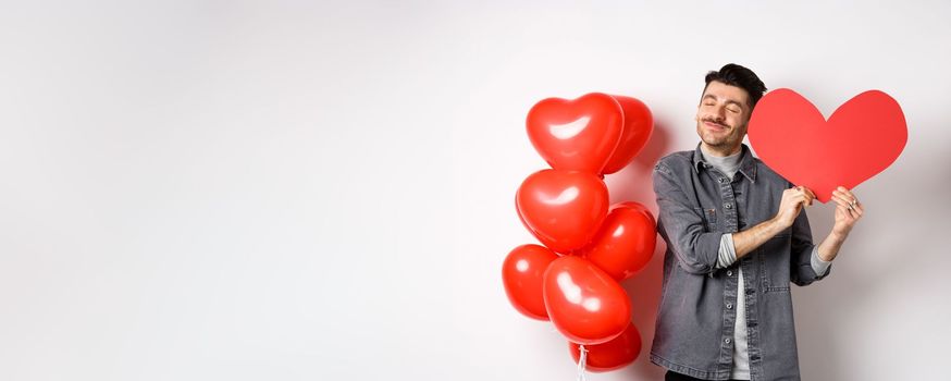 Romantic guy celebrating Valentines day, hugging big red heart card from lover and smiling happy, being in love, standing on white background near red balloons.