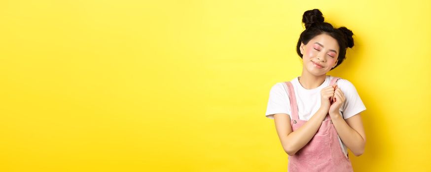 Cute asian girl with makeup, close eyes and remember beautiful moment, holding hands on heart daydreaming, standing on yellow background.