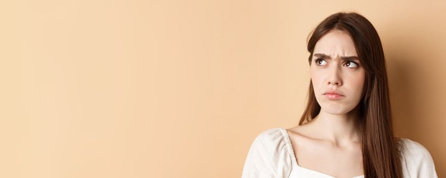 Close up of angry cute girl frowning, looking aside displeased, stare at bad thing, standing on beige background.