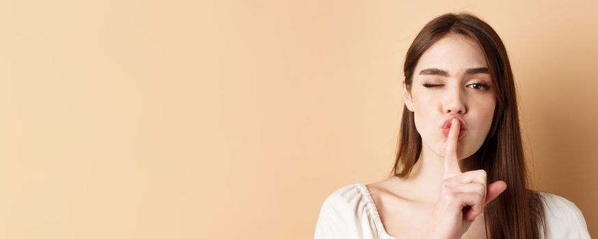 Close up of attractive woman shushing at camera, hiding secret, making hush sign and winking, show taboo sign, standing on beige background.