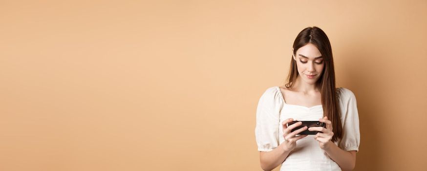 Pretty girl watching videos on smartphone, holding mobile phone horizontally and looking at screen, standing on beige background.