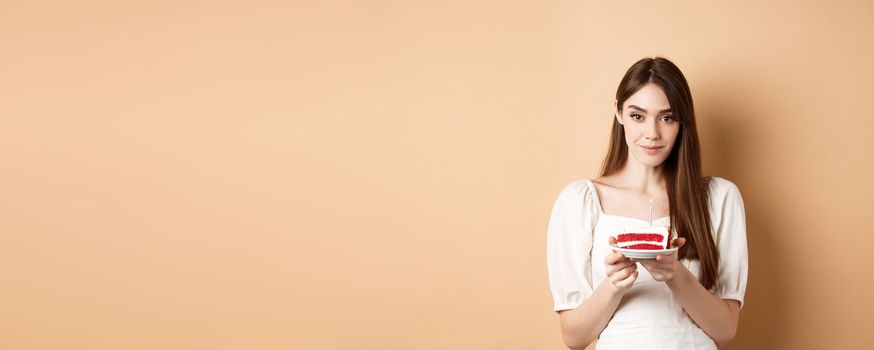 Beautiful birthday girl holding cake with candle, making wish, celebrating b-day on beige background.