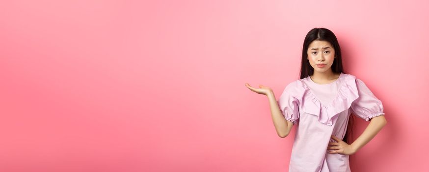 So what, who cares. Indifferent and skeptical asian girl raising arm and look unbothered at camera, dont understand whats wrong, standing against pink background.