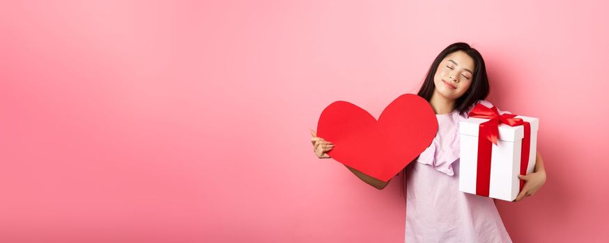 Valentines day concept. Romantic and tender asian teen girl smiling, close eyes and standing dreamy with lover gifts, holding present and big red heart cutout, pink background.