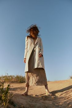 a woman in a fashionable jacket and a long skirt poses standing on the sand against the background of a clear sky. High quality photo