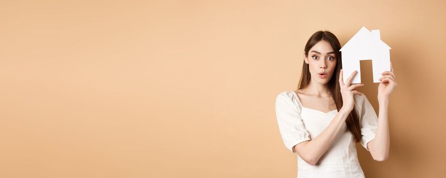 Real estate. Excited young woman showing house cutout and looking at camera, renting property, standing on beige background.