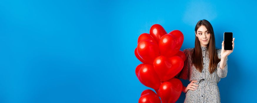 Valentines day. Pretty smiling woman in dress showing empty smartphone screen, standing near romantic balloons, blue background.