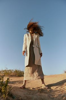a woman in a fashionable jacket and a long skirt poses standing on the sand against the background of a clear sky covering her face with her hair in motion. High quality photo