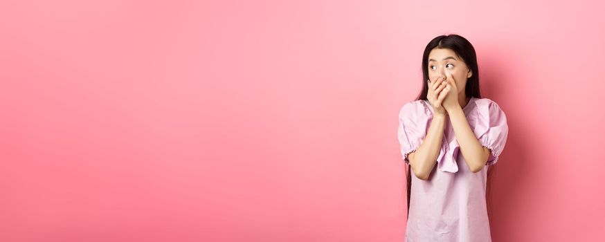Shocked and startled asian woman looking aside at logo, covering mouth with hands speechless, standing in dress on pink background.