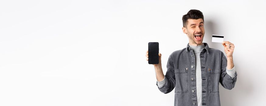 Cheerful handsome guy showing good deal, empty smartphone screen and plastic credit card, winking at you and smiling, recommending offer, white background.