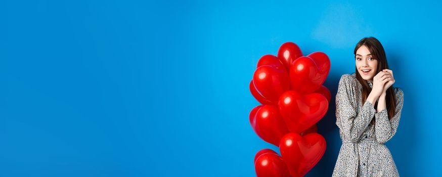 Valentines day. Lovely girlfriend ind ress smiling happy at camera, celebrating lovers holiday, standing near heart balloons, blue background.