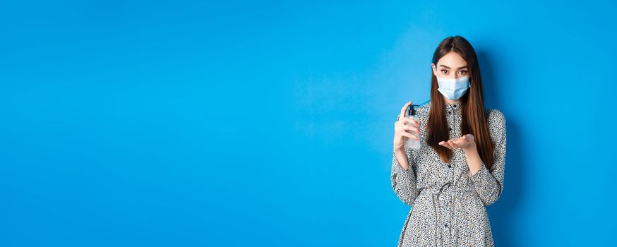 Covid-19, social distancing and healthcare concept. Beautiful girl in medical mask using hand saniziter, holding antiseptic bottle, standing on blue background.