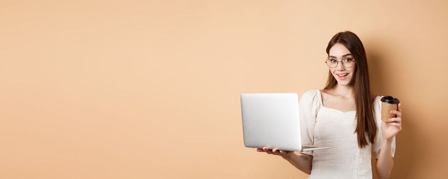 Smiling girl in glasses working on laptop and drinking coffee takeaway. Freelancer using computer at work, standing on beige background.