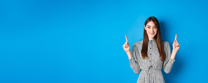 Cute young woman with long hair and vintage dress, smiling and pointing fingers up, advertising on blue background.