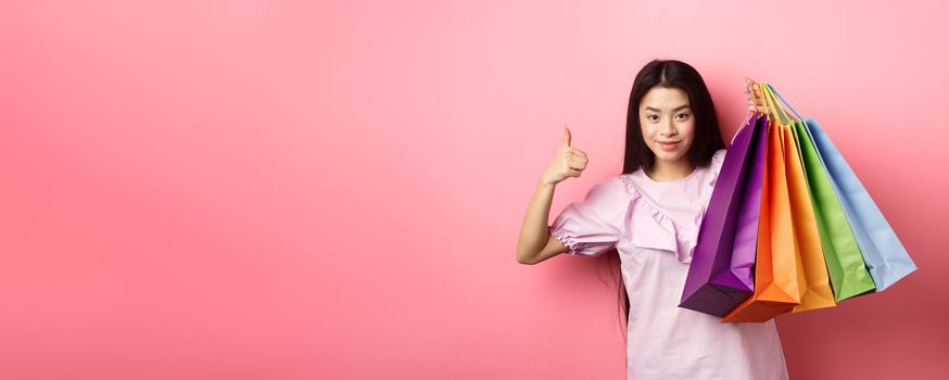 Shopping. Stylish girl showing colorful paper bags and thumbs-up, recommend store discounts, standing against pink background.