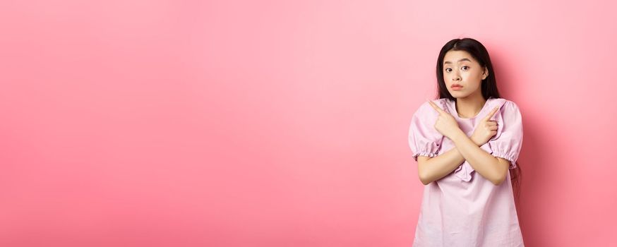 Clueless cute asian girl pointing sideways and look confused, cant make decision, standing hesitant against pink background.