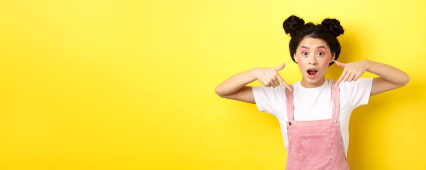 Excited teenage asian girl with beauty makeup, pointing fingers down and open mouth fascinated, standing happy on yellow background.
