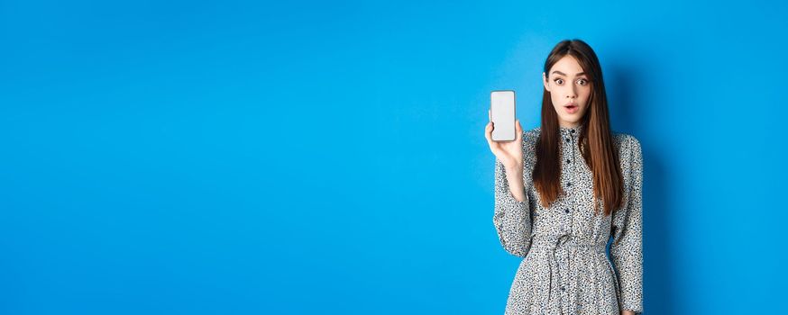 Excited young woman in dress, gasping amazed, showing empty smartphone screen and look impressed at camera, blue background.
