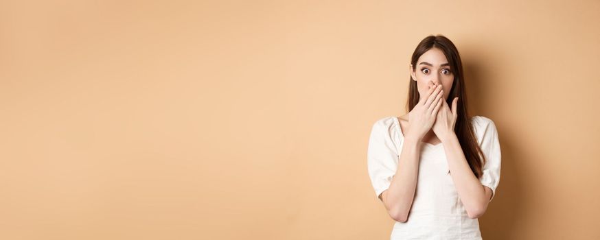 Shocked woman covering mouth with hands and staring startled at camera, standing speechless from gossips on beige background.
