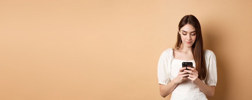 Pretty woman texting on smartphone cell phone, reading screen and smiling, standing on beige background.