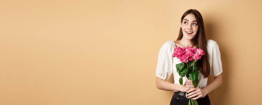 Happy valentines day. Dreamy smiling woman receive bouquet of pink roses and looking aside with love, standing on beige background.