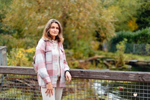 Portrait of cute young woman in casual wear in autumn, standing on bridge against background of an autumn Park and river. Pretty female walking in Park in golden fall. Copy space. smiling girl in the park standing on wooden bridge and looking at the camera in autumn season